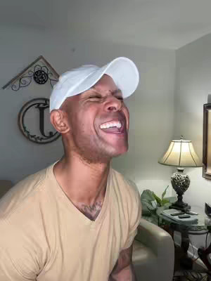 A man in a white baseball cap is laughing with his mouth wide open. He is wearing a tan v-neck shirt. The background includes a tan wall, a beige sofa, a lamp, decor, and part of a glass table.