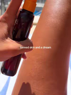 The cover image shows a close-up view of a person's leg being sprayed with a tanning product. The person is holding the product in their left hand and spraying it onto their tanned skin. The background is blurred, but appears to be near water, with blue and white stripes barely visible. The text overlay reads 'tanned skin and a dream'.