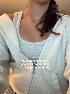 The image shows a person from the chest up, wearing a light grey tank top and a white zip-up hoodie. The person's dark ponytail is visible. Text overlayed on the image reads, "me watching my man serve the Lord and fulfill God's purpose for his life:"
