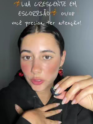 A woman with dark hair and fair skin is in a selfie-style video. She wears a black hoodie and red earrings. Text overlayed at the top reads about a crescent moon in Scorpio with an announcement of the date. The woman is talking and holding a black earbud in her hand.