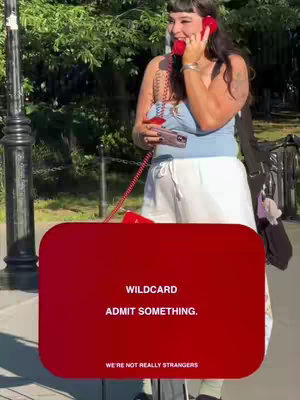 A woman is on a phone call outdoors in a park setting. She is holding a red rotary phone, smiling and looking off to her side. A red rectangle overlay shows the words "WILDCARD ADMIT SOMETHING." with the tagline "WE'RE NOT REALLY STRANGERS".
