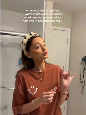 A woman is in a bathroom. She is facing forward, her gaze directed slightly upward. She's holding a lip product with her right hand in front of her face, the product is open with her lips seemingly parted . She's wearing a loose fitting burnt orange T-shirt, an off-white headband with a small black design, and some delicate jewelry.
