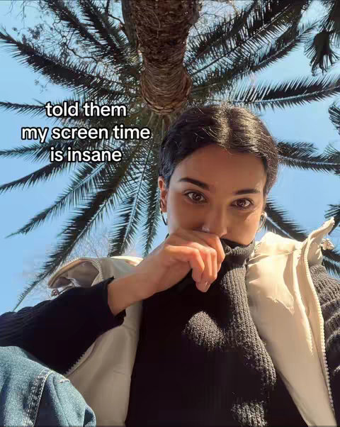 A selfie with a low-angle perspective shows a young woman partially covering her face with her hand. She is wearing a black turtleneck and a white vest, with a palm tree and blue sky forming the background. The text overlay reads, "told them my screen time is insane".