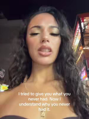 A selfie-style shot featuring a woman with long, dark, wavy hair. Her upper body is visible, and she's looking directly at the camera. The background is blurry but suggests an urban setting with subtle lighting. Text overlayed on the image reads a reflective quote. The overall composition is a close-up, focusing on the woman's face and the overlaid text.