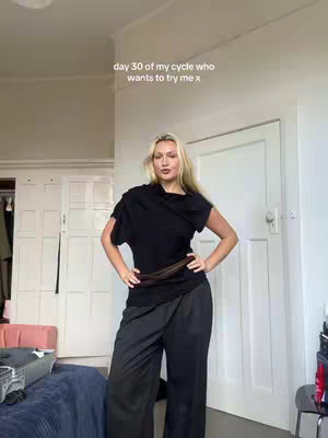 A woman stands in a room wearing a black wrap top and dark pants. The background includes a door and a glimpse of a closet. Text overlay states 'day 30 of my cycle who wants to try me x'.