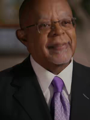 The image presents a close-up of an older Black man wearing glasses and a suit. He has a neatly trimmed beard and a white collared shirt with a purple tie. The background is out of focus, suggesting a studio or indoor setting with diffused lighting. The man's expression is that of a slightly open-mouthed smile, creating a friendly and inviting impression.