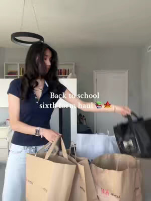 A person stands holding multiple shopping bags, likely filled with school supplies or clothing. The person is partially visible from the shoulders up, wearing a dark blue collared shirt and light-colored jeans. The background shows a room with a neutral color scheme and a hanging circular light fixture. Text overlayed reads "Back to school sixth form haul" with a book and leaf emojis. The person appears to be in their room.