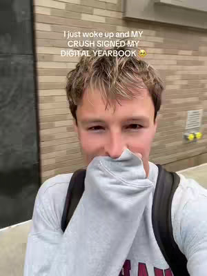 The cover image shows a young man with short, light brown hair. He is holding a gray fabric up to the lower part of his face, covering his nose and mouth. His expression suggests a mix of surprise and happiness. He wears a white long-sleeved shirt and a black backpack. The background features a wall made of rectangular, beige bricks, with a hint of metal on the right. There is text overlaid on the image at the top, written in white.