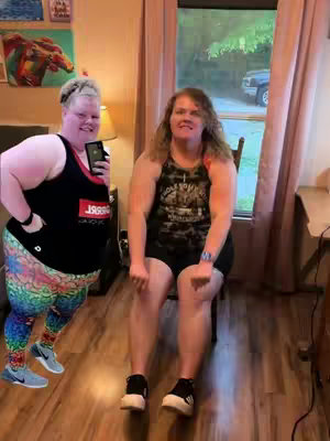 The cover image shows two women in a room setting. One woman is in the foreground on the left wearing a black tank top and colorful printed leggings, holding a phone, and smiling. The second woman is seated on a chair on the right, dressed in black shorts and a top. They are both in what appears to be the middle of exercise. The background includes a window with a view of trees and the outside, and a partially visible door and artwork on the wall. The overall composition gives the impression of a fitness tutorial or workout routine.