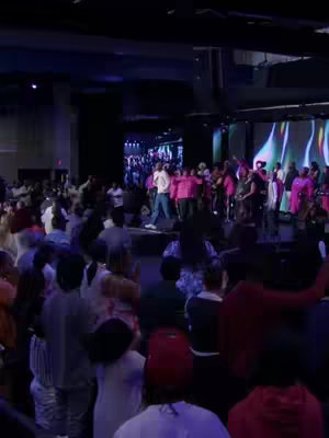 A wide, slightly low-angle shot captures a large, modern auditorium filled with a diverse crowd of people. The stage is brightly lit, with a large screen displaying colorful graphics. A group of people on the stage, presumably a band or choir, are gathered near a central figure dressed in a white outfit, possibly leading the group. The audience members are actively engaged, with many raising their hands, creating an atmosphere of energy and participation. The interior is lit with a combination of stage lights and ambient lighting, creating a vibrant and engaging visual setting.