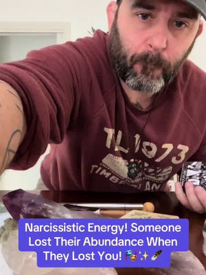 A man with a beard is looking directly at the camera. The background has some objects on a surface and a purple/white rock. Overlay text is on a purple background.