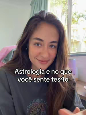 A woman with long, dark brown hair is centered in a selfie-style shot. She's looking at the camera with a slight smile. Text overlay in white in a black box stating "Astrologia e no que você sentie tes4o". The background is slightly blurred, showing indoor setting with a window and natural lighting.