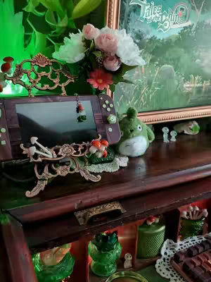 A cozy interior scene featuring a Nintendo Switch console on a wooden shelf, decorated with floral arrangements and small figurines. The game console is set in the foreground with a clear display, flanked by a stuffed Totoro-like creature and a framed picture. The background includes ornate details, with soft lighting enhancing the warm and inviting aesthetic.