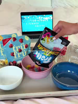 A woman is preparing a movie night setup on a bed. On a wooden tray there are several open bowls, including one filled with colorful candies, and a bag of 'Glumchen' gummy worms. There are also snack bags, a can of soda, glasses, and a laptop showing the 'The Summer I Turned Pretty' title.