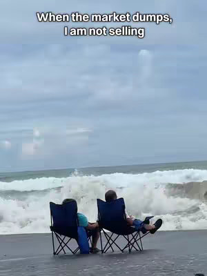 Two people in beach chairs sit on a dark sand beach, gazing out at the ocean. A large wave crashes behind them. The sky above is overcast.