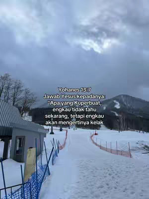 The image shows a snowy outdoor scene. A text overlay provides a bible verse in Indonesian. In the background, there is a mountain range with visible ski slopes, partly obscured by the cloudy sky. A building and a blue fence are visible in the lower left corner.