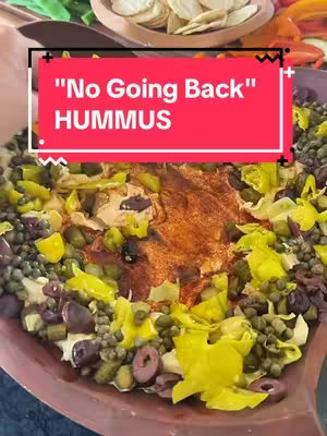 A close-up shot of a hummus dish with a reddish-brown center, surrounded by various toppings like capers, olives, pickles, and pepperoncini, displayed in a brown bowl. A rectangular, red text overlay with white text that reads "'No Going Back' HUMMUS" is placed on top of the dish.