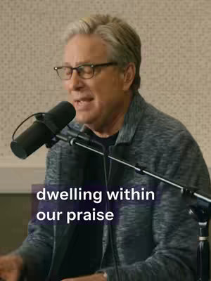 The image shows an older man with gray hair and glasses speaking into a microphone. The background is a textured wall, and overlaid text that reads 'dwelling within our praise'.