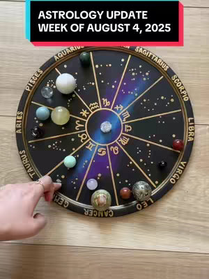 A close-up shot of a circular astrology chart made of wood and decorated with star signs, symbols, and various colorful gemstone spheres arranged across its surface. There is a hand with a ring on the finger interacting with the chart. The background is a light wooden surface.