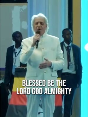 The image shows a man in a white suit speaking into a microphone. Behind him are two men in suits. Text overlayed at the bottom reads "BLESSED BE THE LORD GOD ALMIGHTY". The background appears to be at a religious event with flags visible.