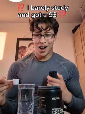 A young man with glasses, holding a clear glass of water and a container of a supplement. He is smiling and has an open mouth with braces. The background is a room with a picture on the wall.