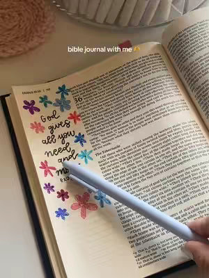 An open Bible rests on a surface, possibly a table or desk. The page is decorated with colorful floral drawings made with gel pens. A light blue pen is also in the image alongside the writing making the image look like a study in bible journaling.