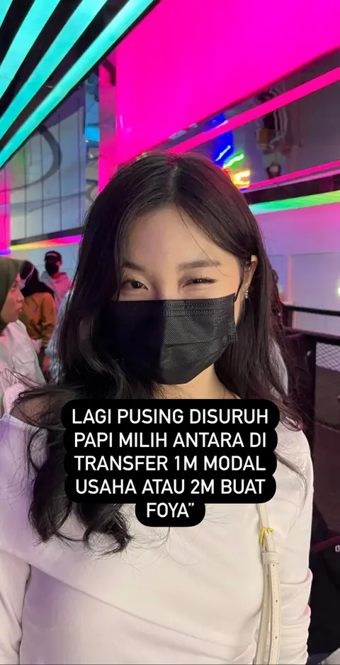 The cover image shows a woman in a mostly light-colored outfit with dark hair and a black face mask. The background features bright pink and blue lights, suggesting an indoor setting. Text overlayed on the image reads in Indonesian. The woman is looking towards the viewer with a slight smile.