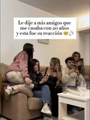 The image shows a group of friends reacting to an announcement. The foreground features a group of four women and a man seated on or near a light brown couch. The hook text is in a white box at the top, reading 'Le dije a mis amigos que me casaba con 20 años y esta fue su reacción'. The women's faces are contorted with shock and expressions of surprise. Their attire is casual. In the background, the setting appears to be a living room or similar indoor space. The overall composition is framed by a warm, well-lit environment.