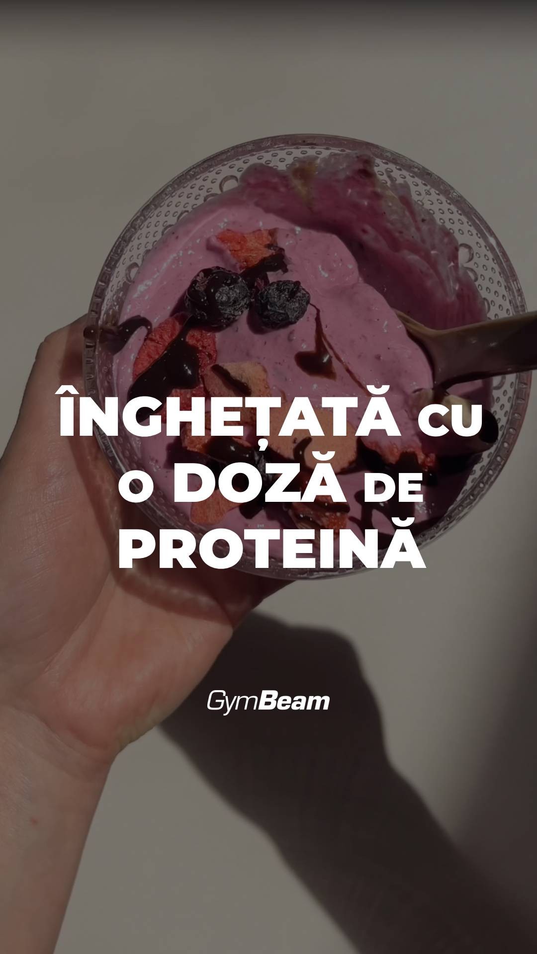 A hand holding a clear glass bowl filled with pink-colored ice cream, topped with blueberries and chocolate sauce. The text overlayed over the image reads: "ÎNGHEȚATĂ CU O DOZĂ DE PROTEINĂ" placed at the center of the image. The GymBeam logo is placed at the bottom. The background is neutral and slightly out of focus.