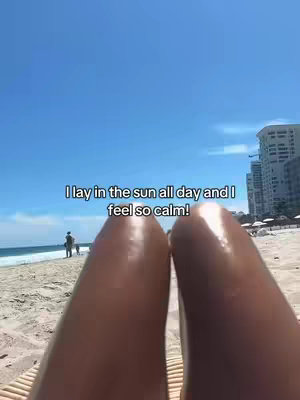 A first-person view of legs, the bright sun, and blue sky on the beach. The legs are in the foreground, and the beach and ocean are in the background. A line of text "I lay in the sun all day and I feel so calm!"  is at the center.