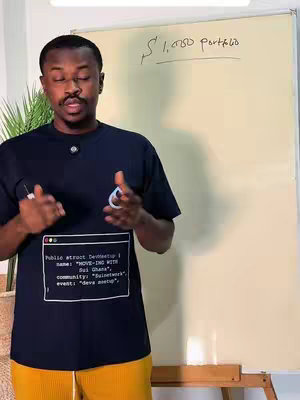 A man stands in front of a whiteboard in a room with a yellowed board. He is gesturing towards it with his hands. He is centered in the shot and is wearing a black t-shirt with design on the front and mustard-colored pants.