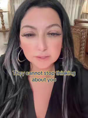 A close-up shot of a woman with long, dark hair with silver streaks. She is wearing large hoop earrings and a black top. Text overlayed on the image reads, 'They cannot stop thinking about you'. The blurred background suggests an indoor setting, possibly a home.