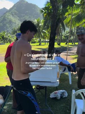A man is standing shirtless with his back to the viewer, leaning toward a white cooler, with a woman in a red shirt and another man in a cap on the left and right, respectively. There's text overlaid in the center. The background includes a mountain, trees, and some other people.