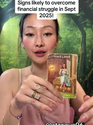 A woman holds up a tarot card with a serene image while looking directly at the camera. The background shows a green backdrop with leafy patterns, and the woman wears light makeup with highlighted cheeks, and jewelry, including large rings. The tarot card features a woman in a blue dress seated near a treasure chest. Overlaying the scene is white text.