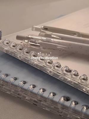 The image showcases a close-up view of study materials. A blue spiral notebook is positioned in the foreground, featuring a clear, plastic binding. Resting atop are two pens and what appears to be a clear, plastic pencil case. The color palette is muted, with soft blues, whites, and translucent elements providing a clean and organized aesthetic. The phrase 'study motivation' is overlaid on the image, indicating the video's thematic direction.