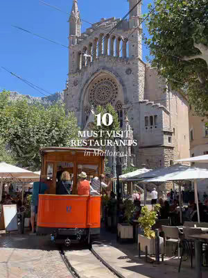 A photo featuring a historic tram moving on rails with passengers in the foreground, a towering stone church in the background under a bright blue sky, and tables and chairs from a cafe. The image is vibrant, capturing a sunny day in a European city.