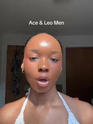 A front-facing shot of a woman looking directly at the camera. She has dark hair, and her skin appears to be a rich brown tone. She has makeup on her face and is wearing a white top. The text overlay reads "Ace & Leo Men". The background is neutral with a glimpse of a wardrobe and door.
