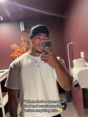 A man with tattoos is holding a phone up to his face, taking a selfie in a bathroom. The background has a pinkish hue. The caption added is a quote about a woman of God wanting marriage.