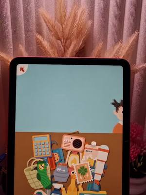 A tablet displaying a colorful game interface. The foreground features various cartoon-style objects, and the background is a soft pink curtain.
