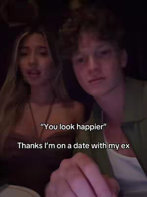 A dimly lit selfie-style shot features a young couple, with a woman on the left and a man on the right. The woman looks towards the camera and is wearing a brown top, her blonde hair falling around her face. The man has short, light-colored hair and is looking off to the side. Text overlays the image, reading "You look happier" and "Thanks I'm on a date with my ex."