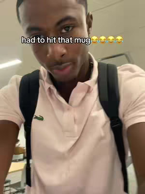 A medium shot of a young Black man standing in a hallway, wearing a light pink polo shirt and a black backpack. He's looking towards the camera with a slight smile and a slightly raised eyebrow, with the text 'had to hit that mug' overlaid at the top.