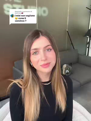 A woman with long blonde and brown hair is looking at the camera with a neutral expression. She's wearing a black top and a delicate necklace. Behind her is a blurred background with soft, neutral tones and a glimpse of a grey couch, a microphone and some set lighting.