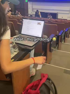 A person is sitting in an auditorium, holding a laptop. The laptop is open and displaying a screen. The person is wearing a white t-shirt and a blue hat. The background is blurred, showing rows of seats and other people.