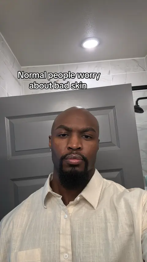 A bald Black man is in a bathroom, looking at the camera with a serious expression. Text overlayed on top reads, "Normal people worry about bad skin."