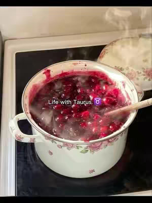 A close-up shot of a pot on a stovetop filled with a boiling, deep red mixture, possibly jam or sauce. Steam rises from the pot, and a wooden spoon rests inside. Text overlayed in the center reads: Life with Taurus with a Taurus zodiac sign symbol. The pot has a floral design.