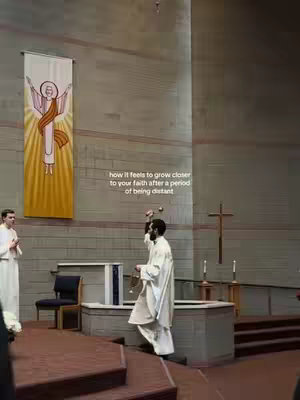 A religious ceremony taking place inside a church. The image is a medium shot with a focus on the altar area. A priest in white robes stands near a table with candles, swinging a censer. Another person in white robes is on the left. Behind them is a tall banner with an illustration. The color scheme is muted, with neutral tones of beige and brown, and a red brick floor adding the primary contrast.