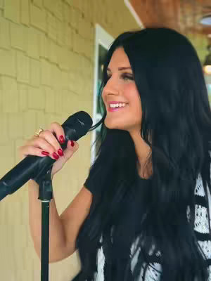 A woman with long, dark hair smiles while holding a microphone. She's wearing a black shirt and a patterned white and black cardigan. The background features a wall with light-colored siding and a partial view of a door.