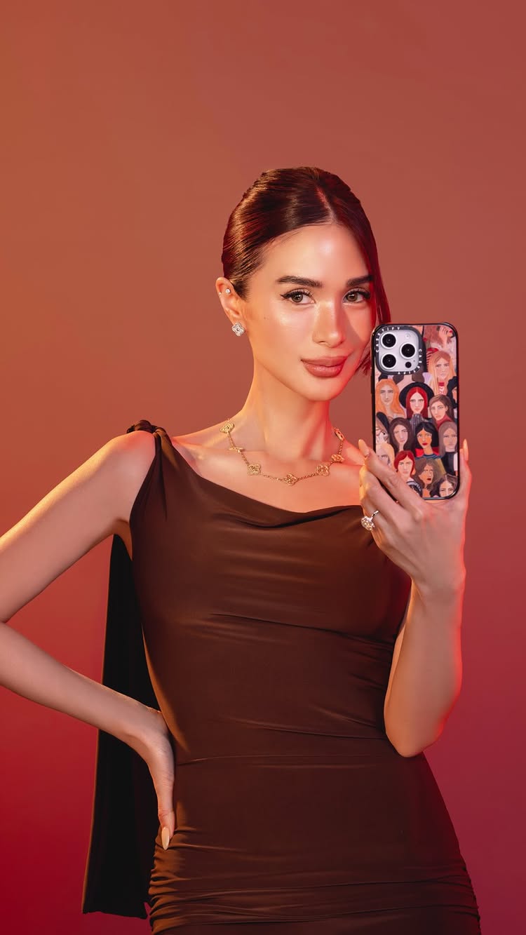A well-lit portrait of a woman holding a phone case. The background is a gradient of warm colors. The woman is in a dark brown dress, with her hair up. She's wearing jewelry and has a phone in her hand, the back of the phone is covered with a illustration of multiple faces. The woman smiles at the camera, her pose is composed.