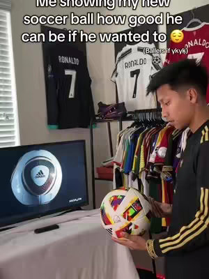 A young man holds a soccer ball in front of a TV screen. The TV shows an image of a soccer ball, similar to the one he's holding. The background includes soccer jerseys hanging on a rack and mounted on the wall