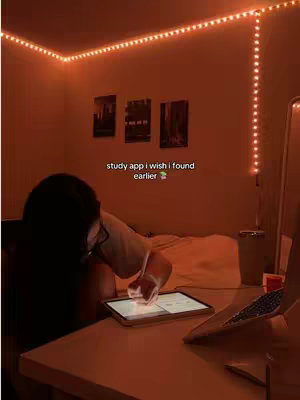 A person is working on a tablet at a desk in a dimly lit room. The room has warm, orange lighting from desk lamps and string lights. Text on the image reads: "study app i wish I found earlier". The background shows a bed with some art prints on the wall. A laptop is visible on the desk, and a tumbler is sitting beside it.
